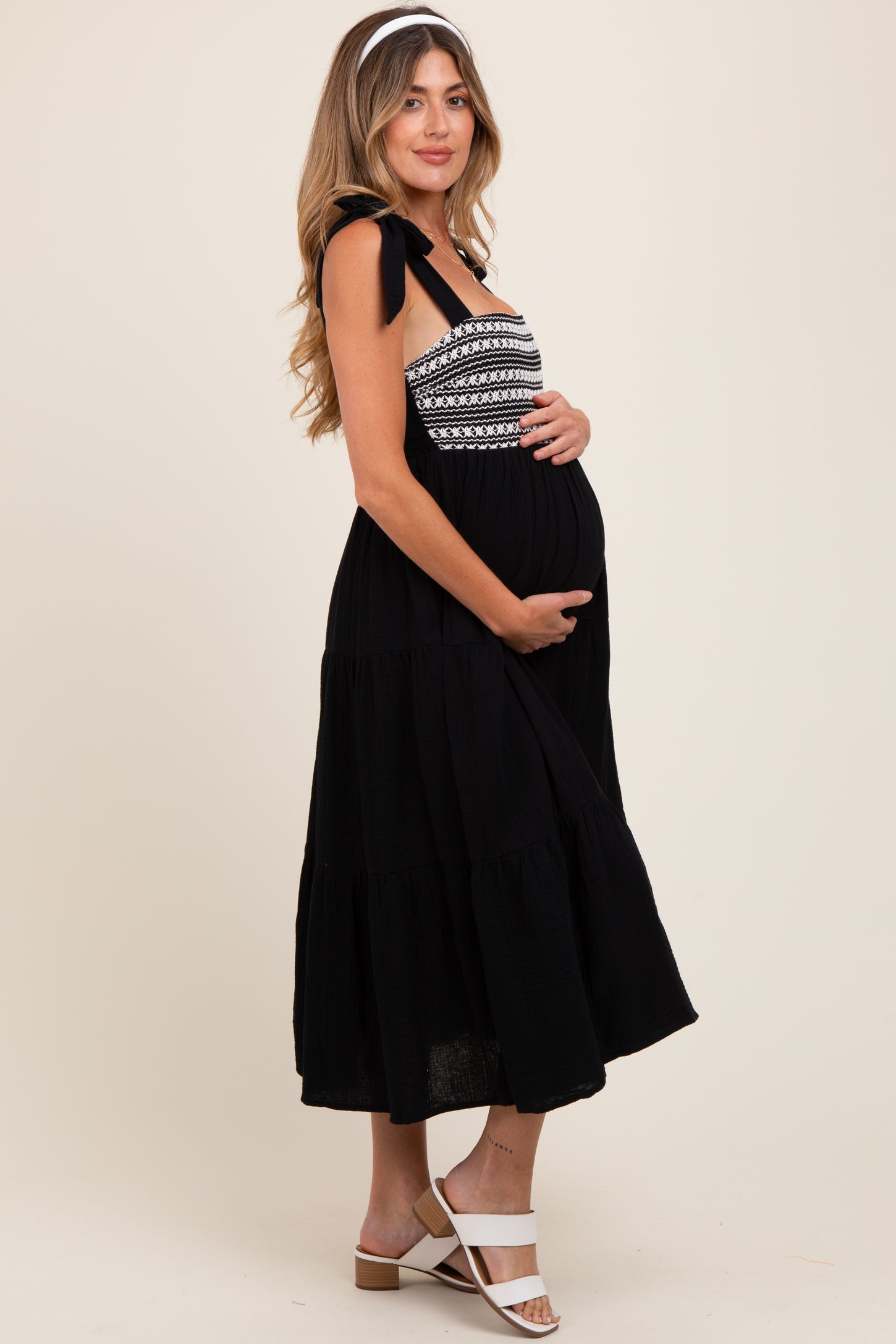 woman wearing a black maternity maxi dress with embroidered bodice and tie straps, holding her baby bump