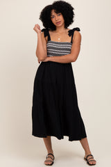 woman wearing a black midi dress with embroidered bodice and tie straps, standing confidently with one hand on her hip
