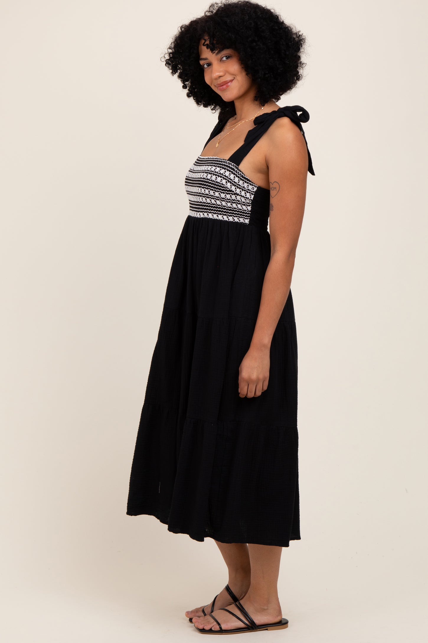 woman wearing a black midi dress with embroidered bodice and tie straps, standing sideways with a relaxed expression