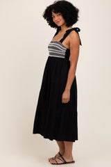 woman wearing a black midi dress with embroidered bodice and tie straps, standing sideways with a relaxed expression