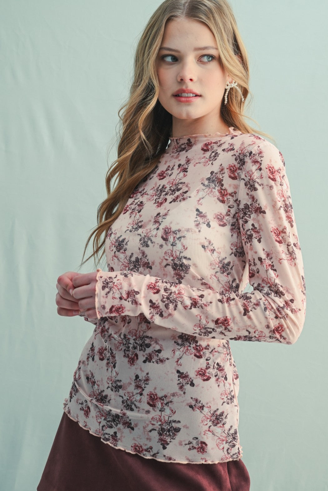 woman wearing a light pink long sleeve top with floral print and ruffled hem, standing against a pale background