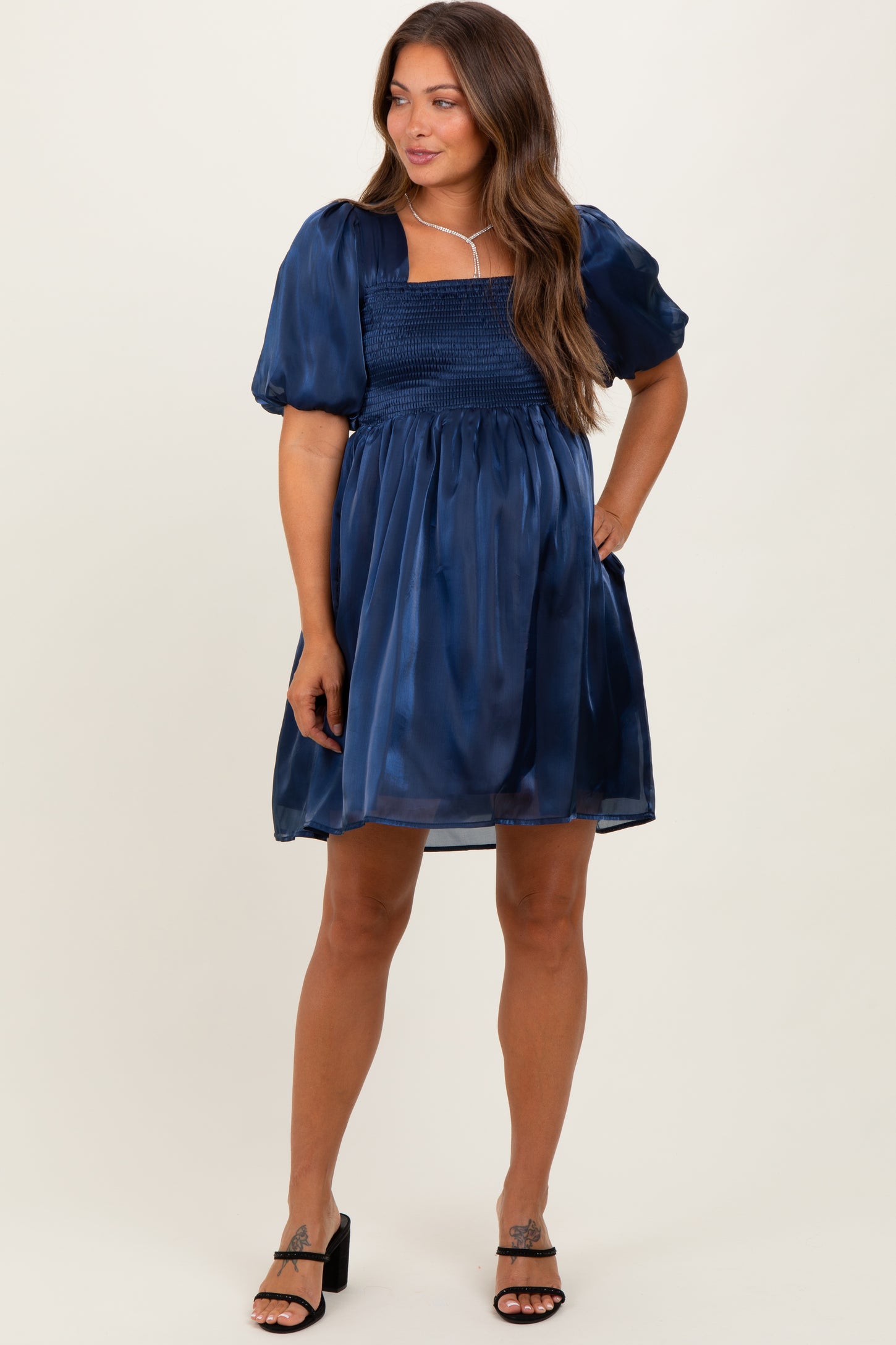woman wearing a navy blue short puff sleeve maternity dress with a smocked bodice and flowing skirt, holding her baby bump