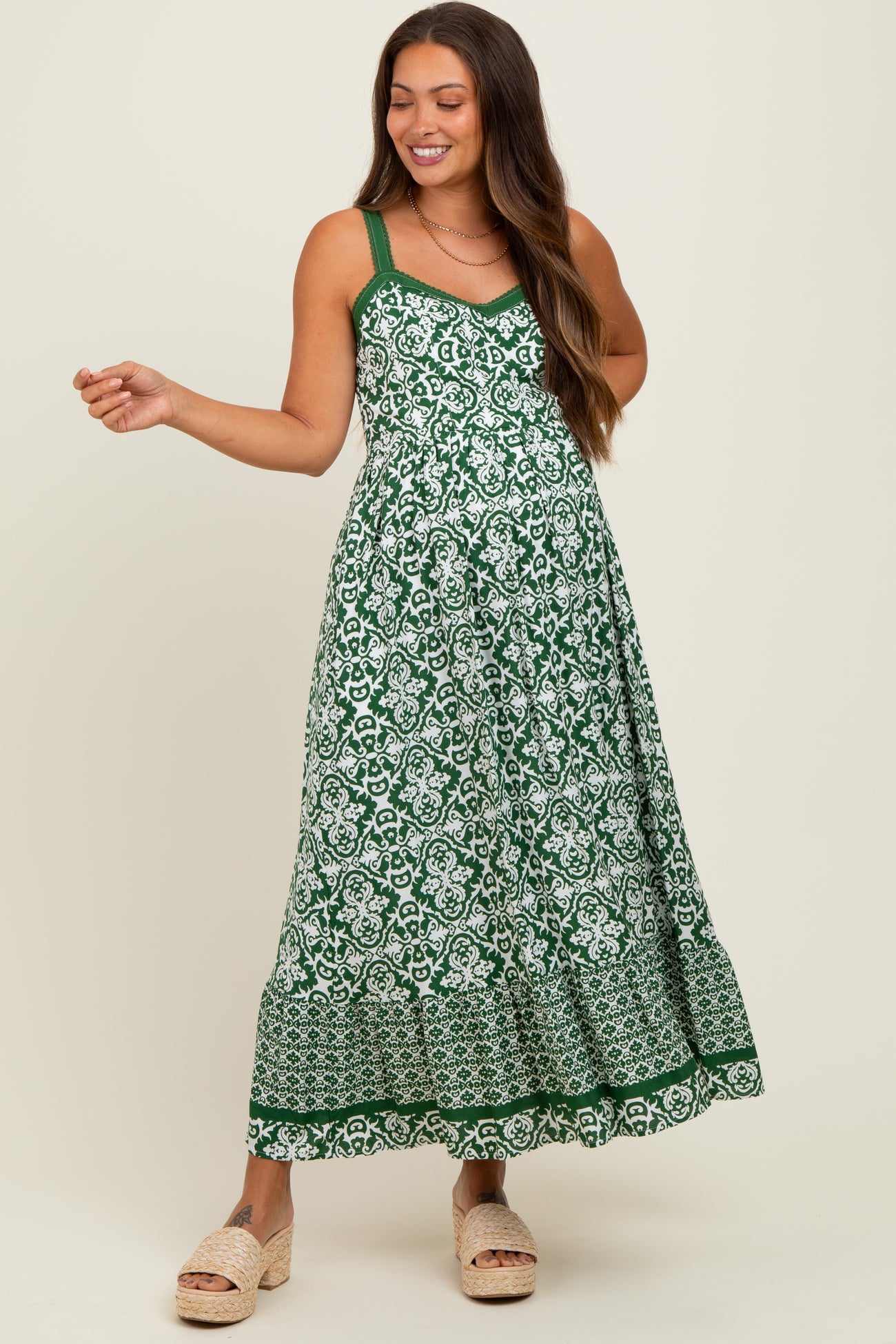 woman wearing a green floral print maternity dress with spaghetti straps and a flowy skirt, smiling and posing