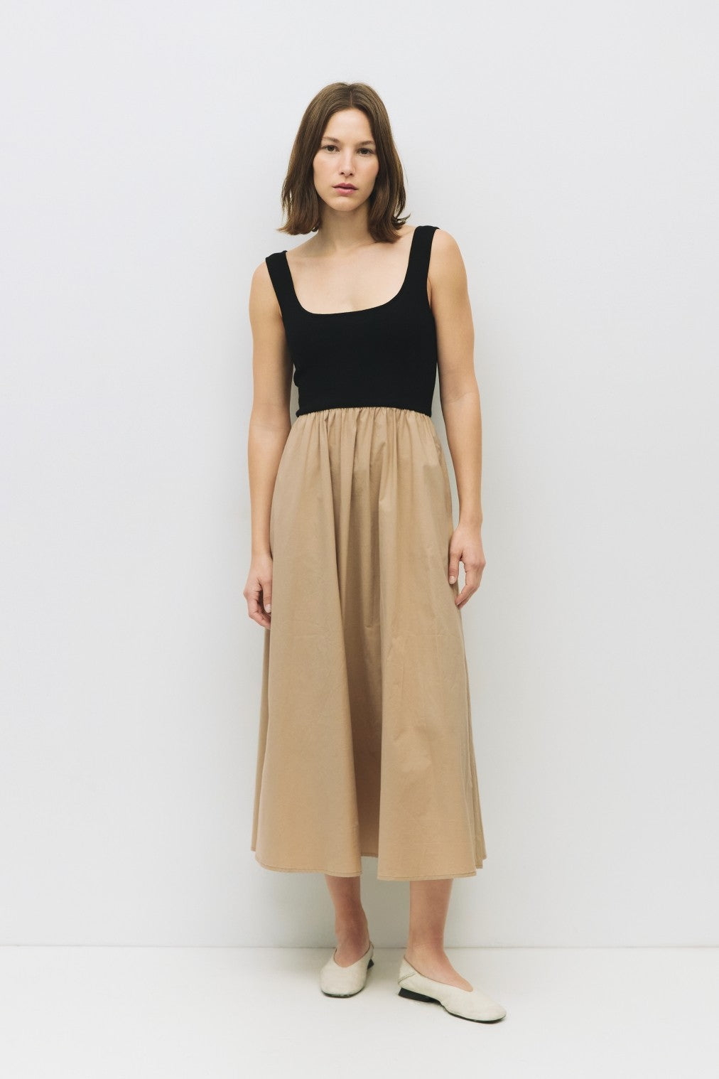 woman wearing a black sleeveless top and a tan midi skirt, standing against a white background