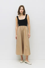 woman wearing a black sleeveless top and a tan midi skirt, standing against a white background