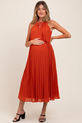 woman wearing an orange pleated maternity dress, holding her baby bump with a chic hairstyle and black heels