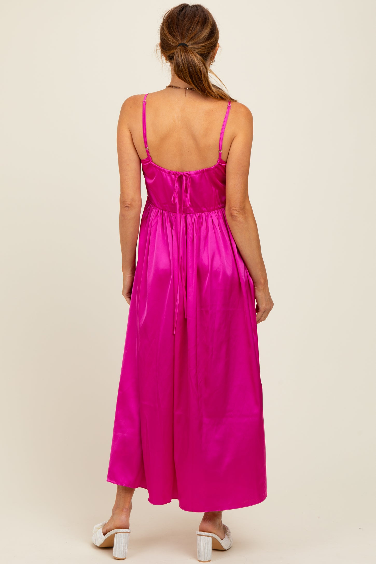 woman wearing a back view of a bright pink satin maxi dress with adjustable straps and a tie closure at the back
