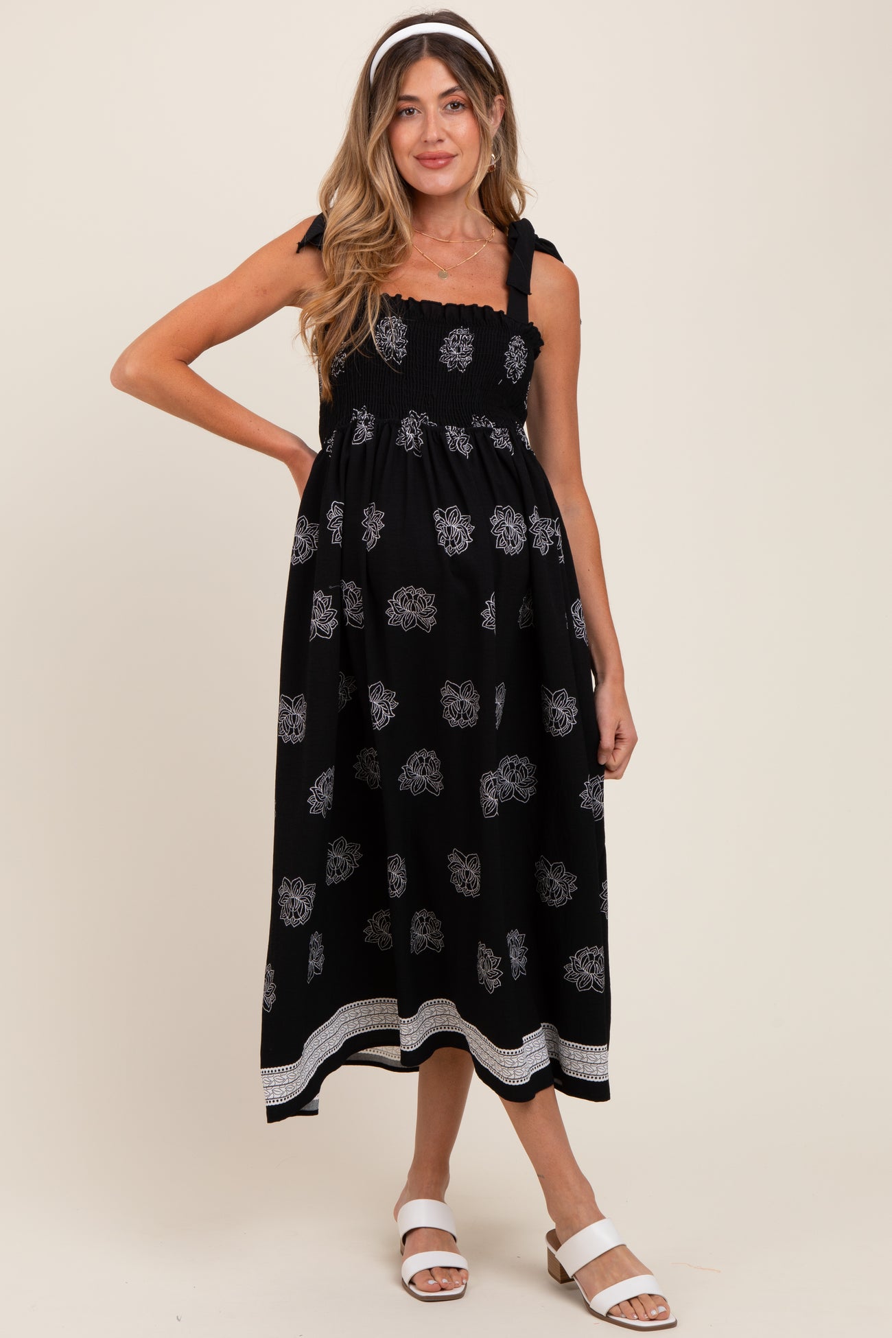 woman wearing a black maternity dress with floral print, smocked bodice, and tie straps, posing with a baby bump visible