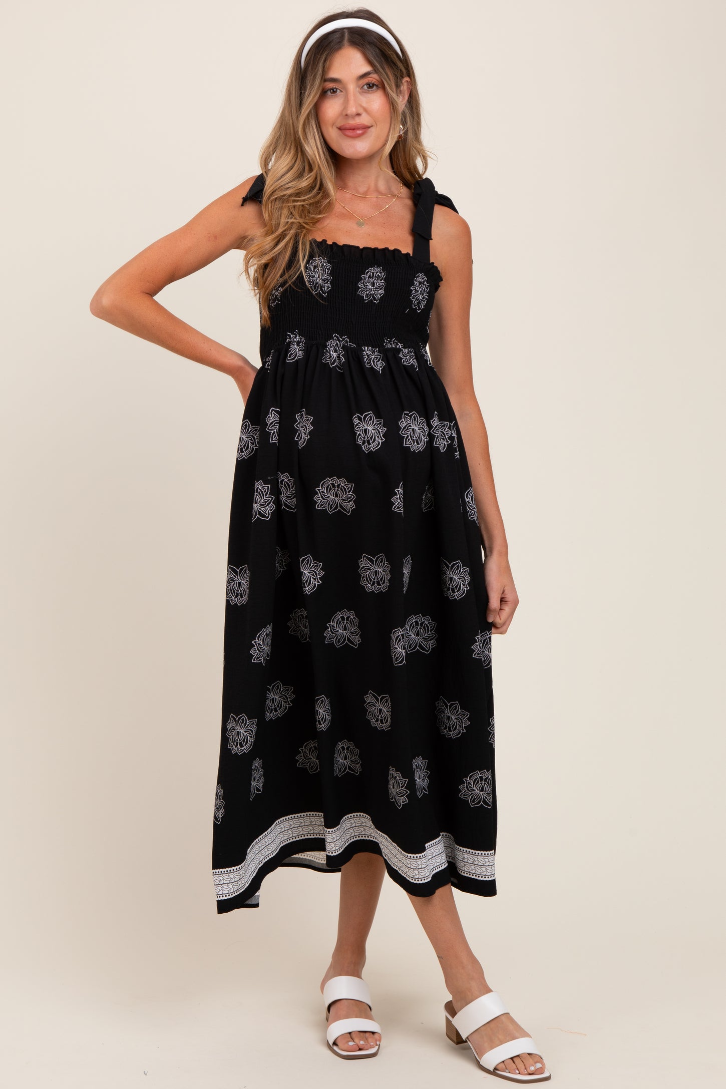 woman wearing a black maternity dress with floral print, smocked bodice, and tie straps, posing with a baby bump visible