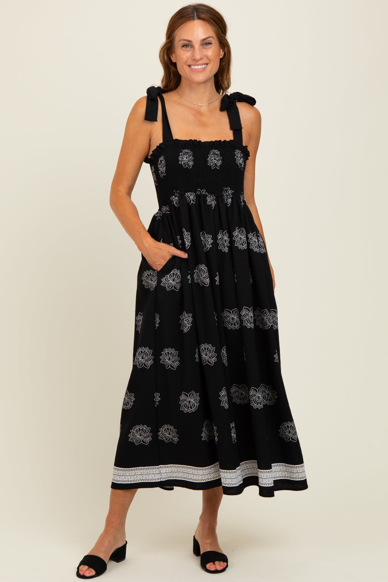 woman wearing a black maxi dress with floral patterns and tie shoulder straps, standing with pockets