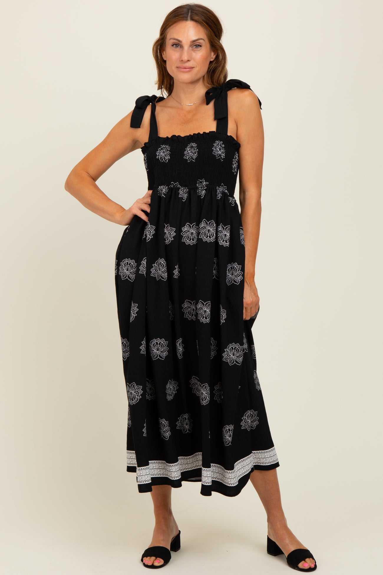 woman wearing a black midi dress with white floral patterns and tie shoulder straps, standing with one hand on her hip