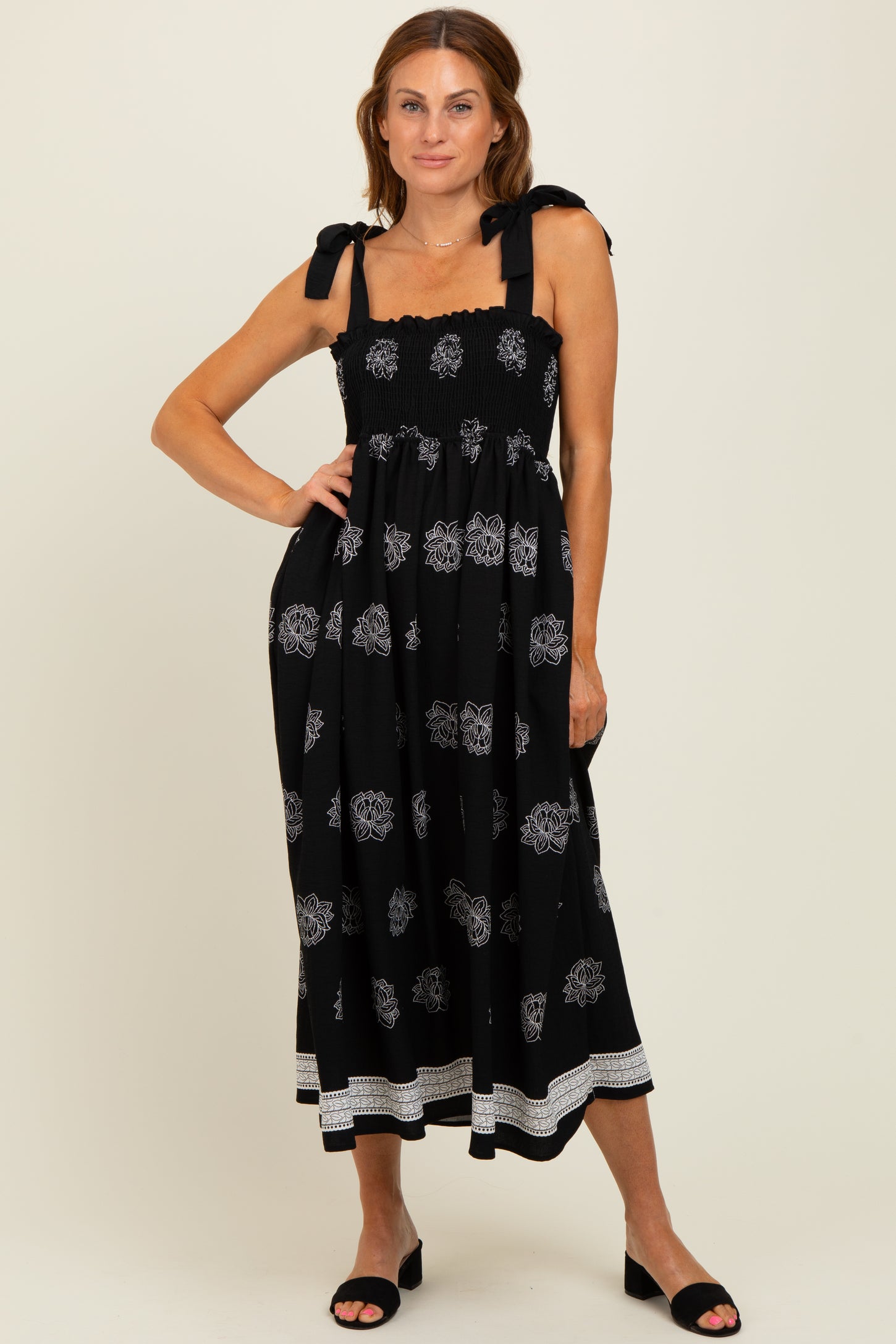 woman wearing a black midi dress with white floral patterns and tie shoulder straps, standing with one hand on her hip
