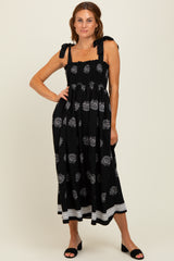 woman wearing a black midi dress with white floral patterns and tie shoulder straps, standing with one hand on her hip