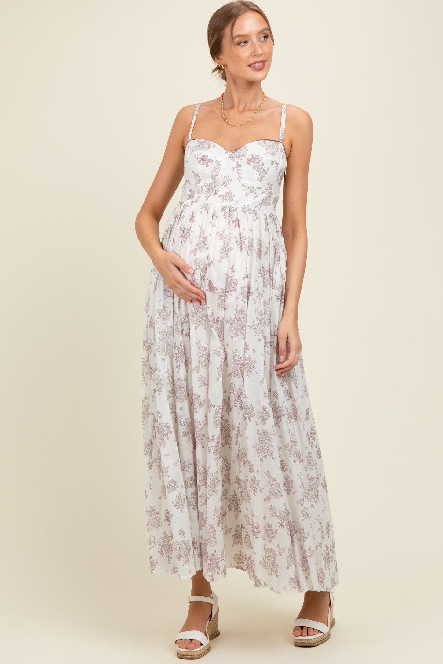 woman wearing a white floral maternity maxi dress with spaghetti straps and holding her baby bump