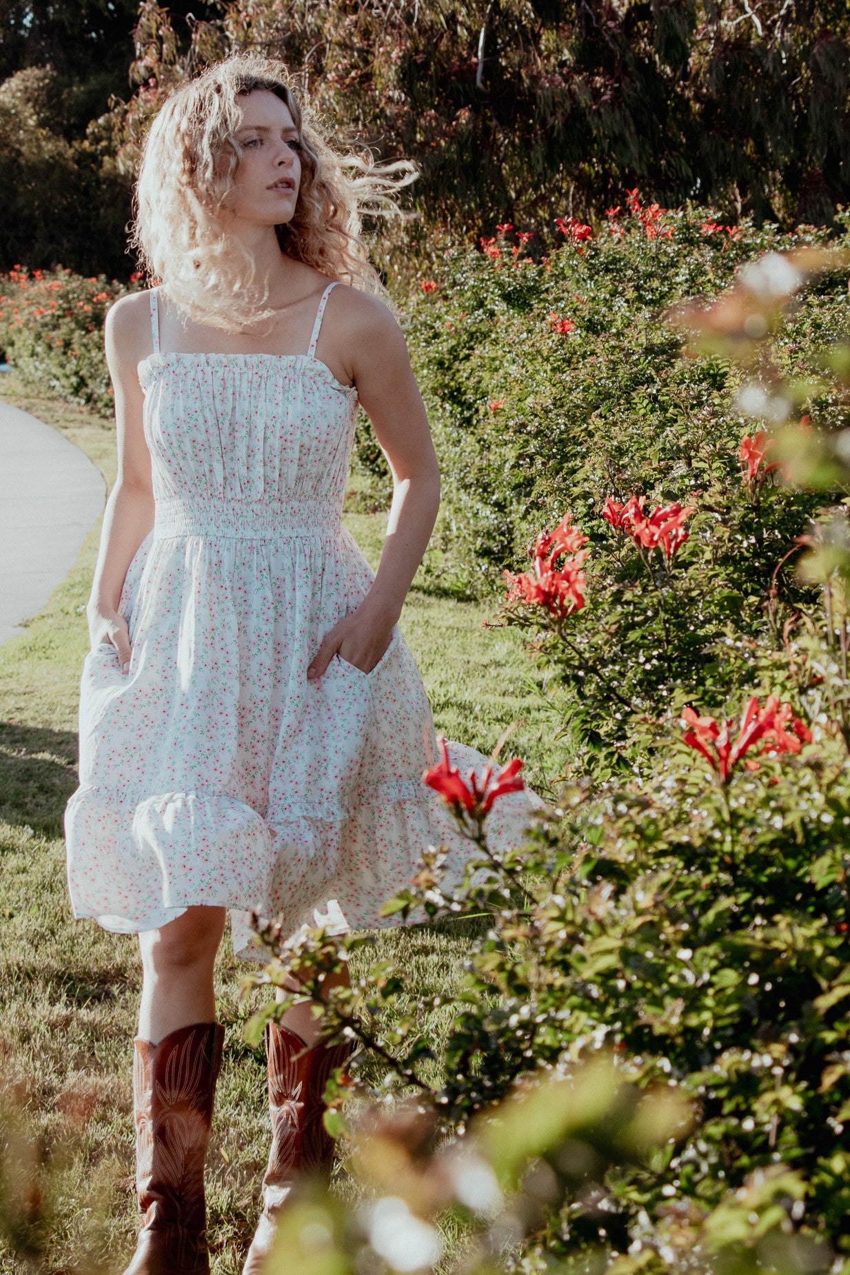 woman wearing a white floral sundress with a smocked bodice and pockets, paired with brown cowboy boots in a garden setting
