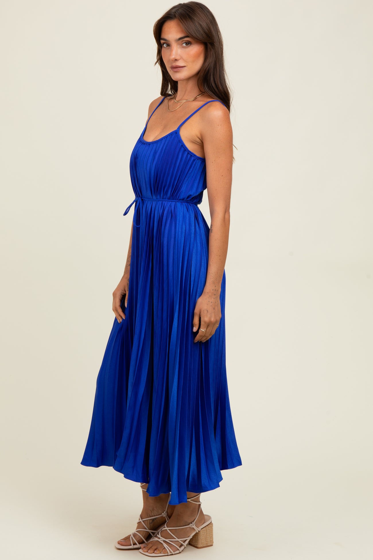 woman wearing a sleeveless royal blue pleated maxi dress with a tie waist and strappy sandals