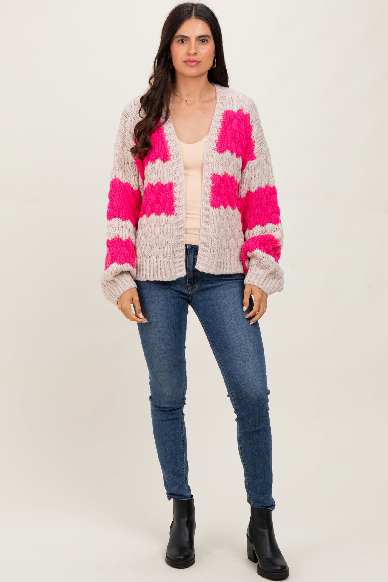 woman wearing a light pink cardigan with bold pink patches over a cream top and blue skinny jeans, standing in black ankle boots