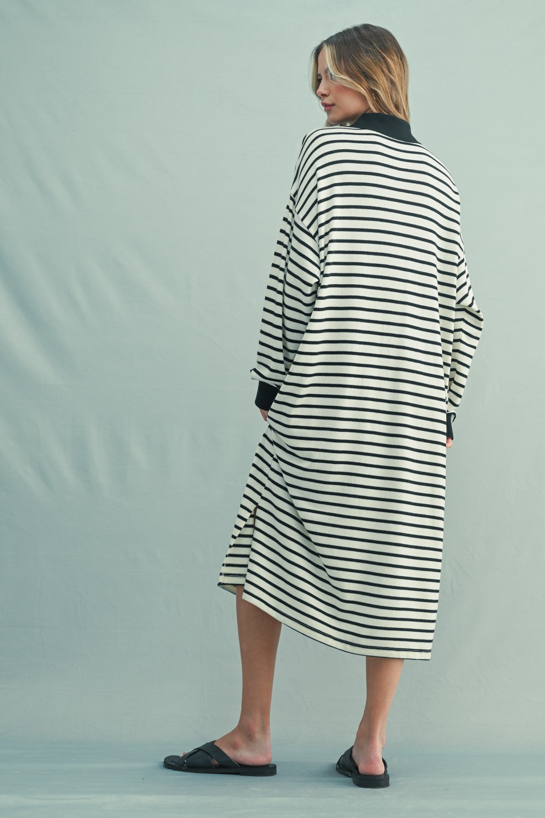 woman wearing a black and white striped oversized dress with long sleeves and side slits, standing with her back turned