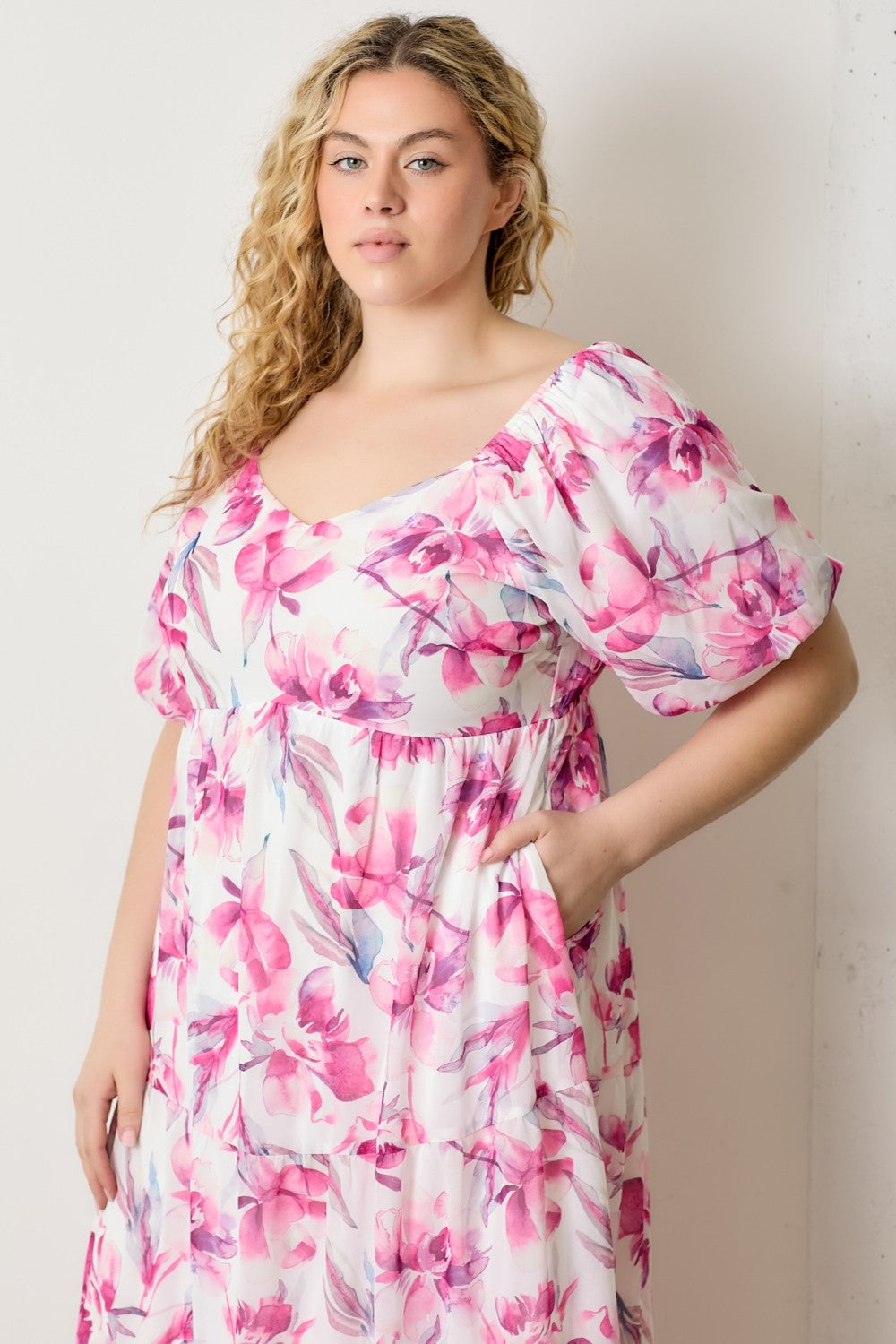 woman wearing a floral plus size dress with pink and white colors and puff sleeves, posed with hands in pockets