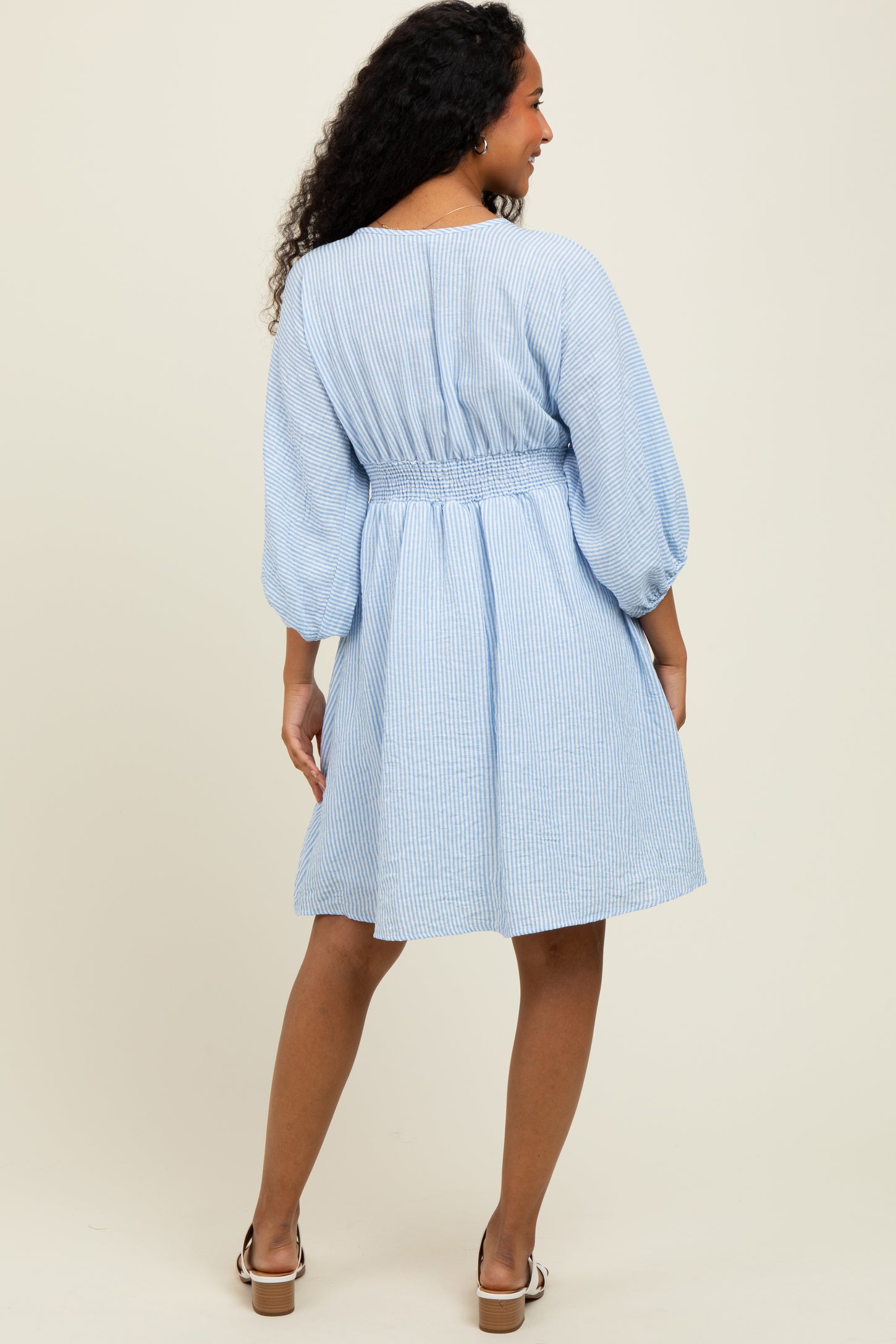 woman wearing a light blue striped short dress with a smocked waist and balloon sleeves, viewed from the back