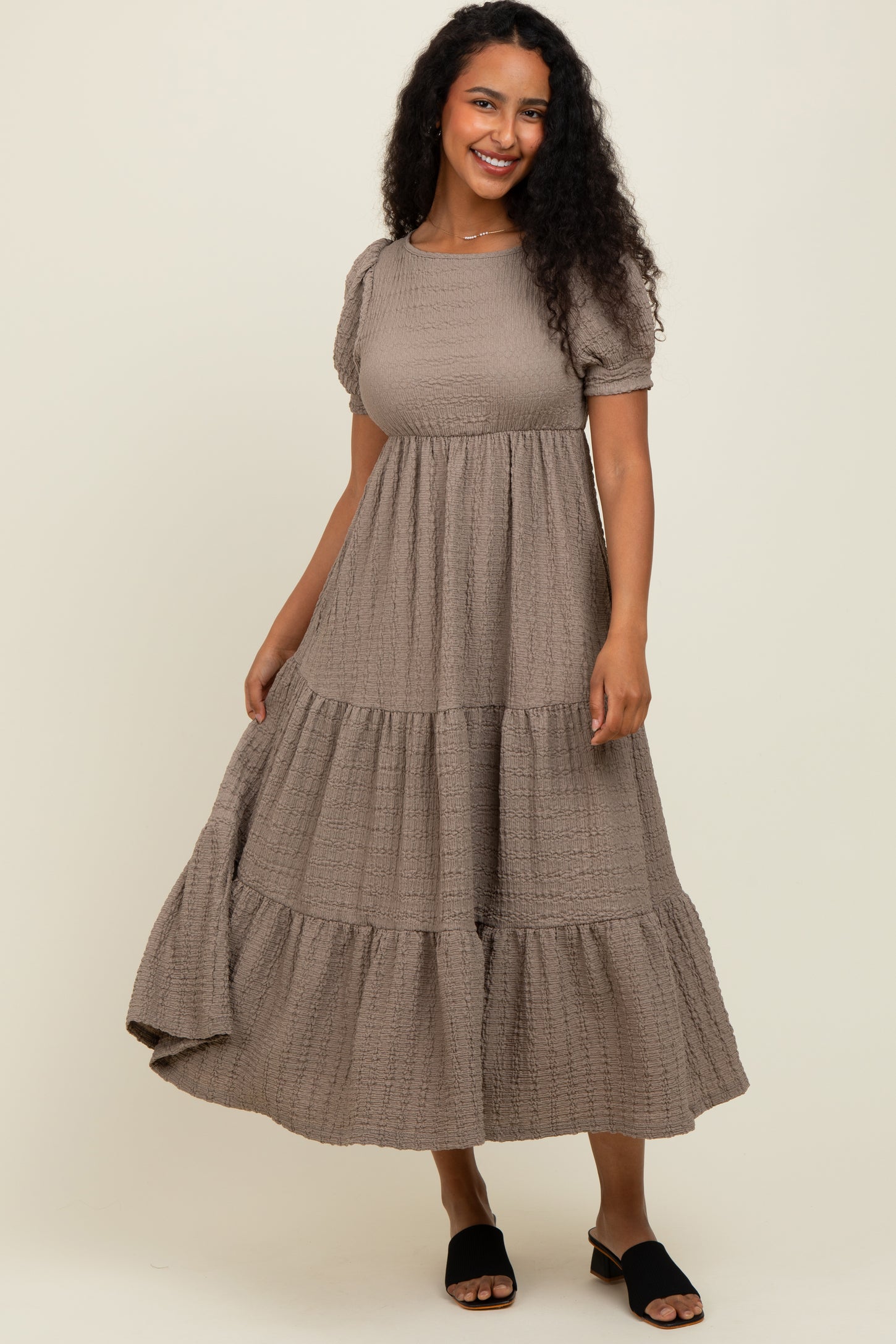 woman wearing a taupe tiered puff sleeve maxi dress with a textured fabric and black slide sandals