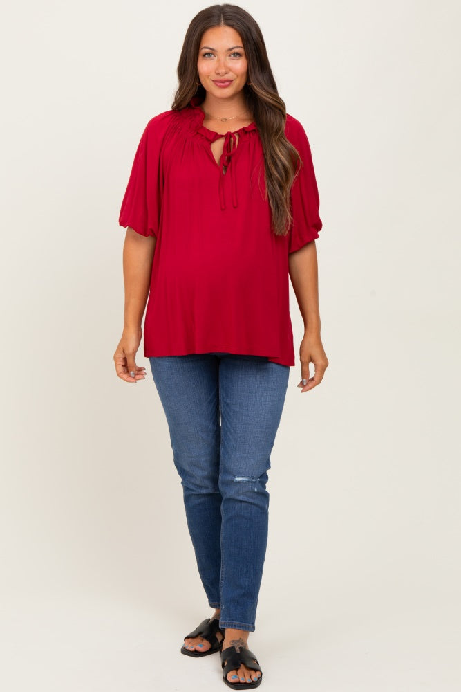 woman wearing a burgundy maternity top with a tie neckline and puff sleeves, paired with blue jeans and black sandals, showing a baby bump