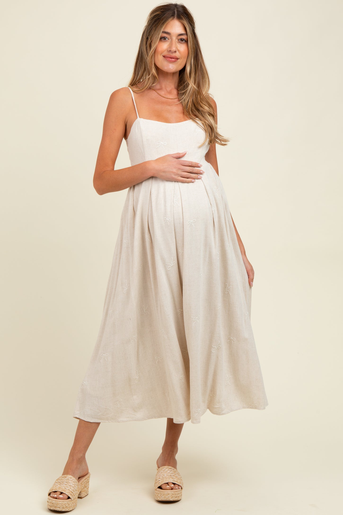 woman wearing a light beige maternity dress with spaghetti straps and a flowy silhouette, holding her baby bump