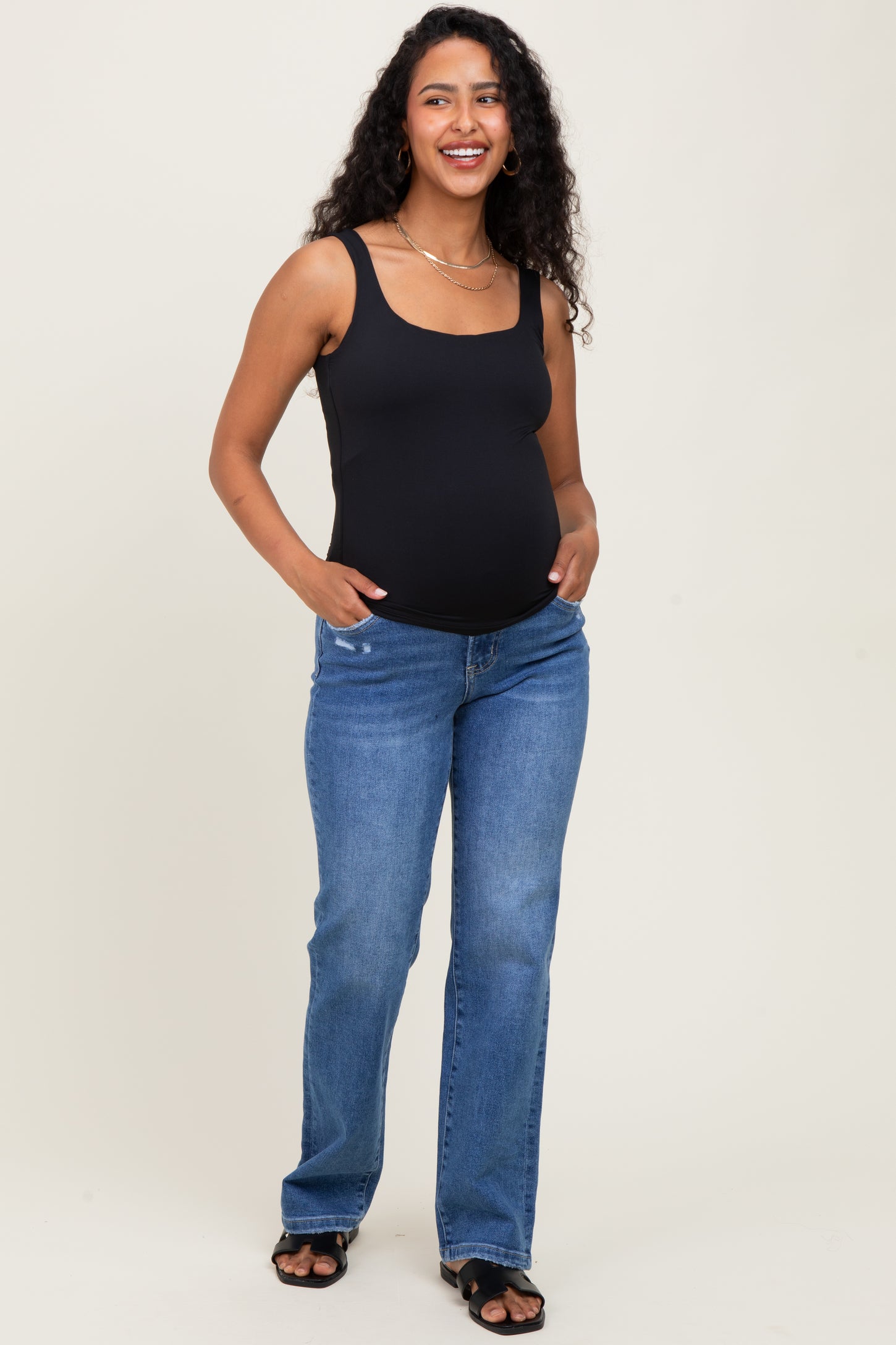 woman wearing a black maternity tank top and blue jeans with a visible baby bump, standing and smiling
