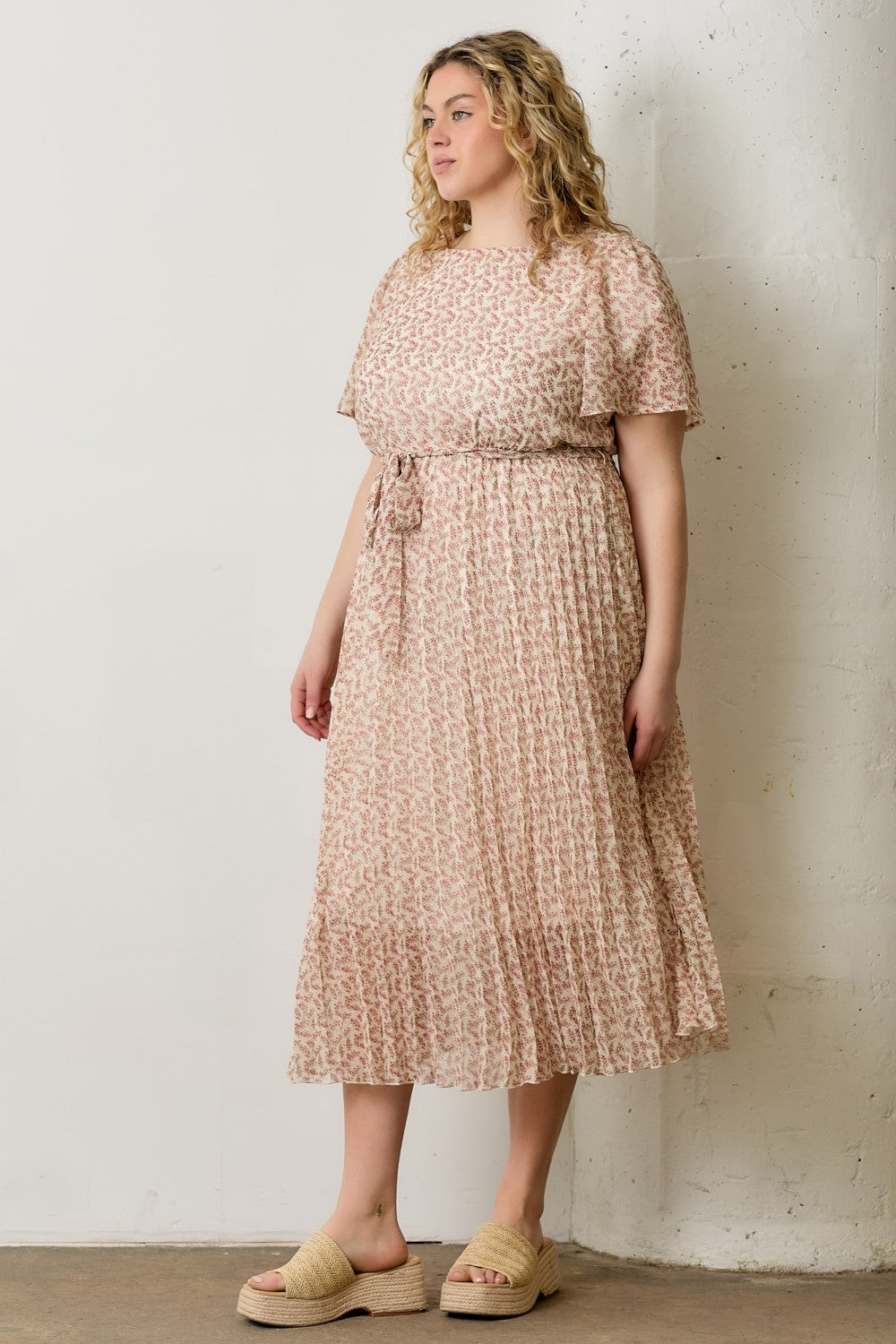 woman wearing a light pink plus size midi dress with a floral pattern and short sleeves, standing against a wall
