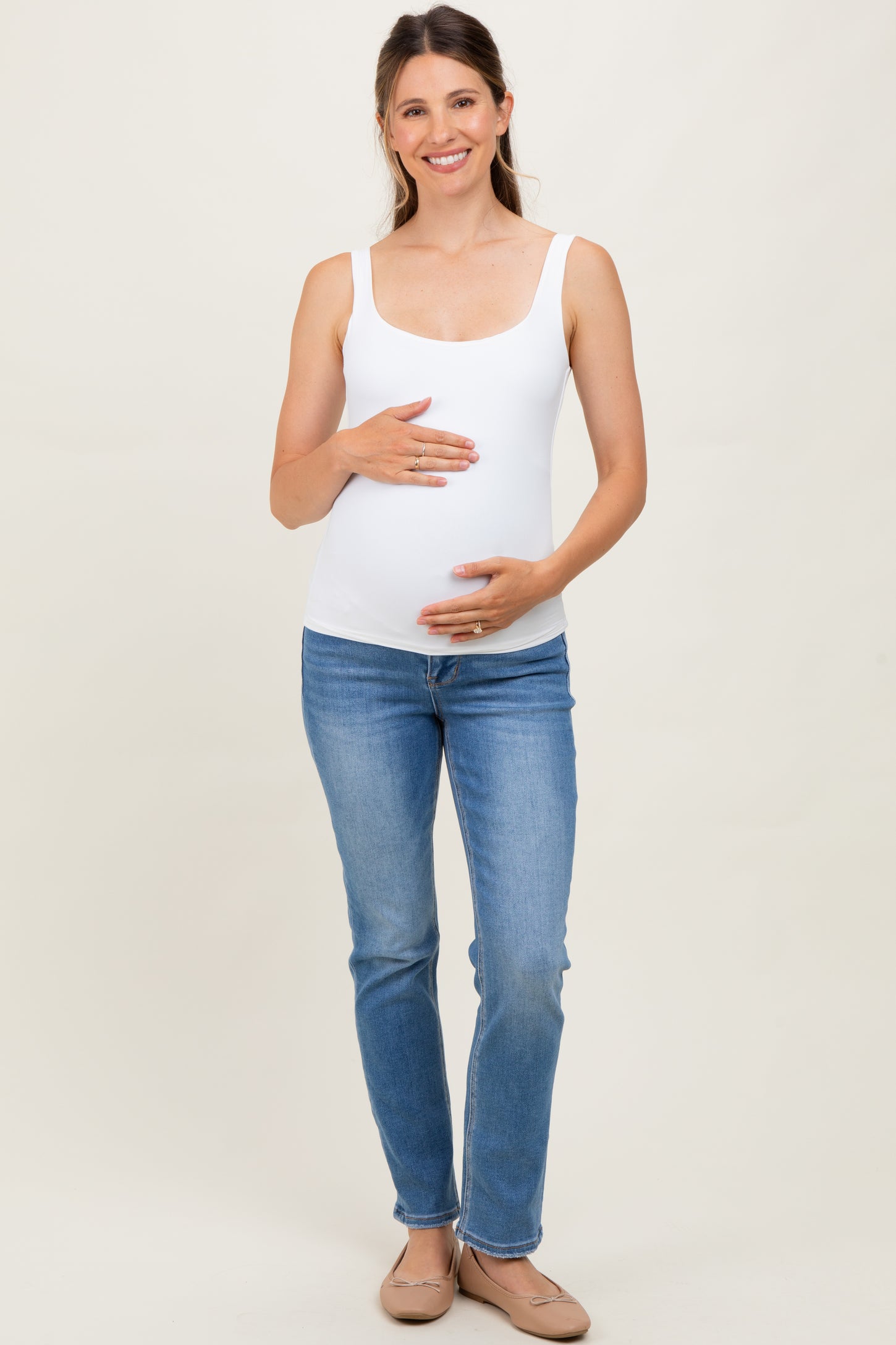 woman wearing a white maternity tank top and blue jeans, holding her baby bump with a smile