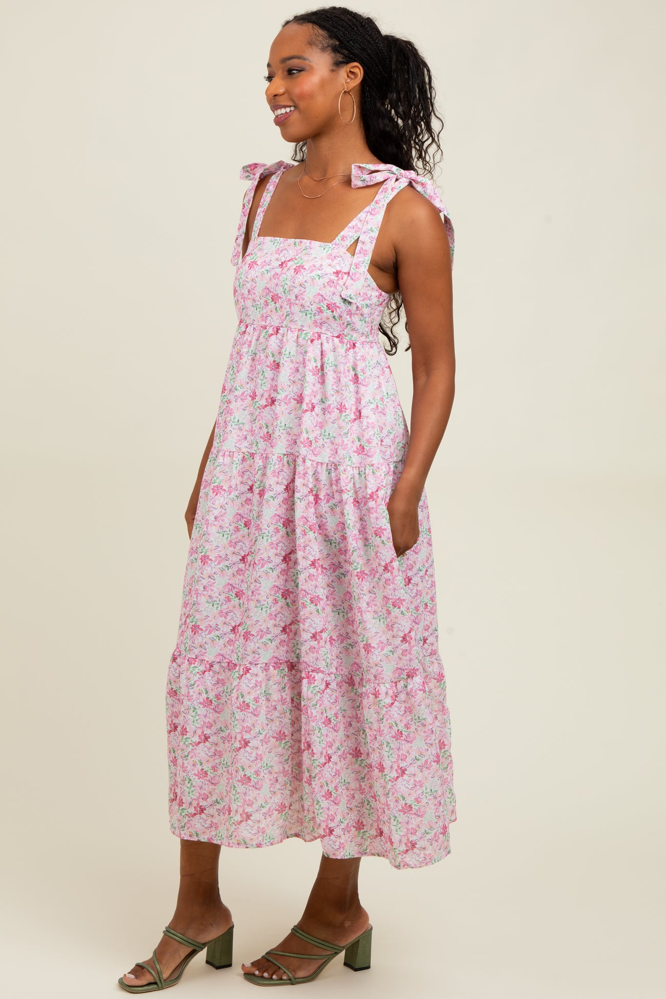 woman wearing a pink floral maxi dress with bow tie straps and pockets, standing sideways with a relaxed pose