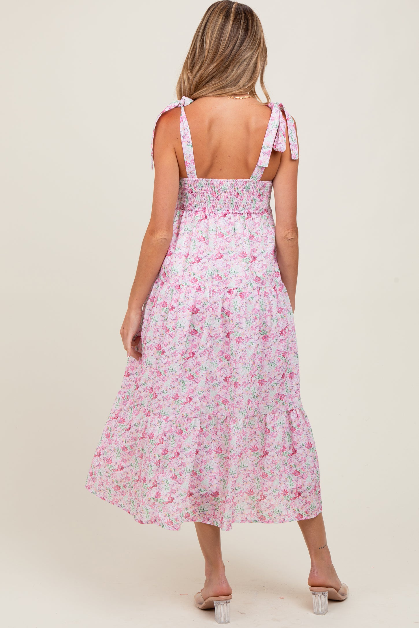 woman wearing a pink floral maternity dress with adjustable straps and a flowy tiered skirt, showing her back and baby bump