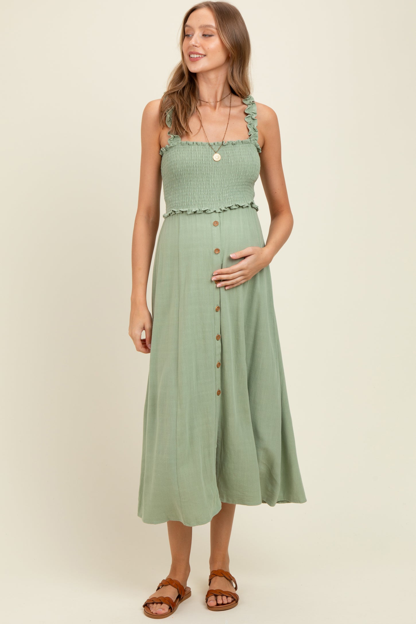 woman wearing a light green maternity dress with ruffled straps, smocked bodice, and button details, holding her baby bump