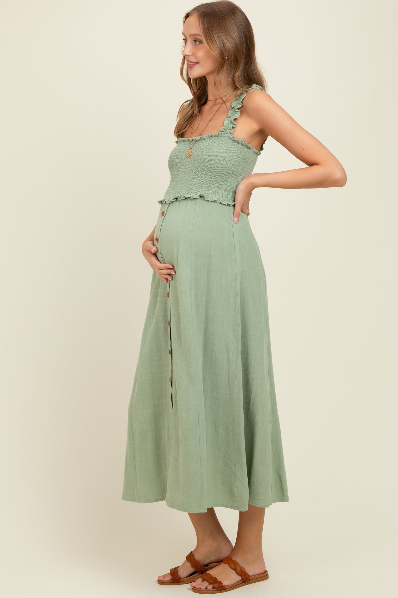 woman wearing a sage green maternity dress with smocked bodice and button details, gently cradling her baby bump