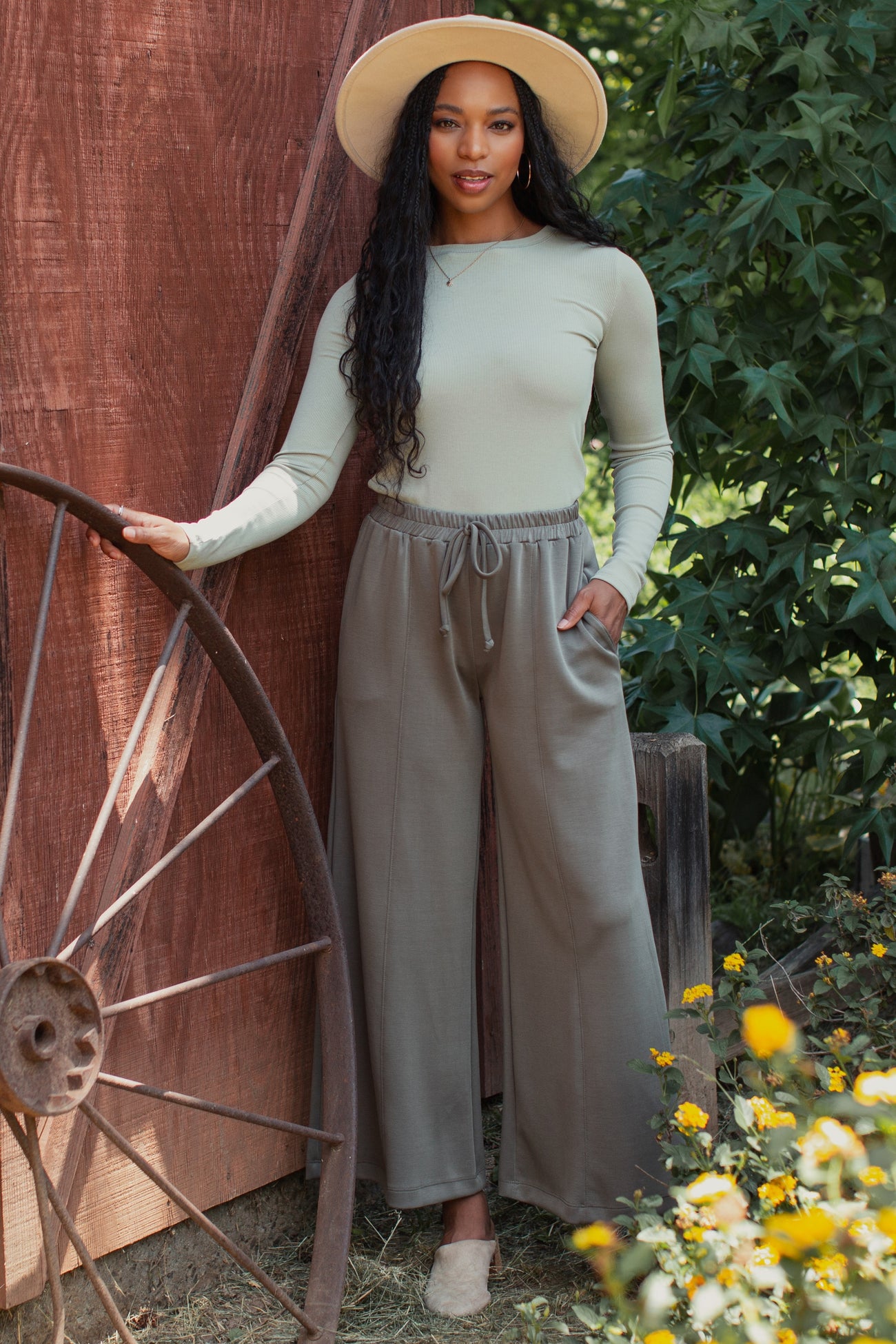 woman wearing a light green long sleeve top and taupe wide-leg pants, posing outdoors with a hat and flower backdrop