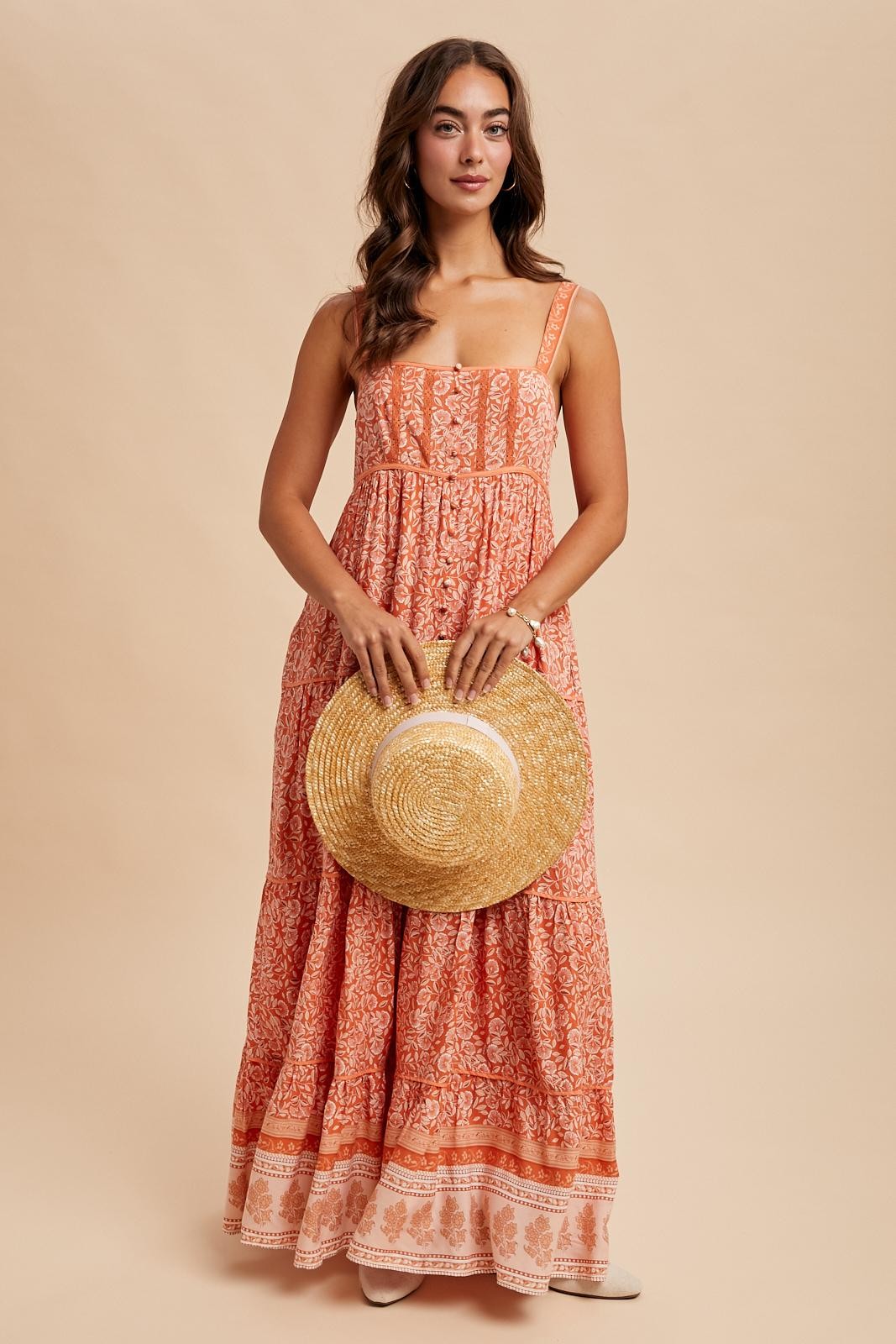woman wearing a rust colored maxi dress with floral patterns and holding a straw hat