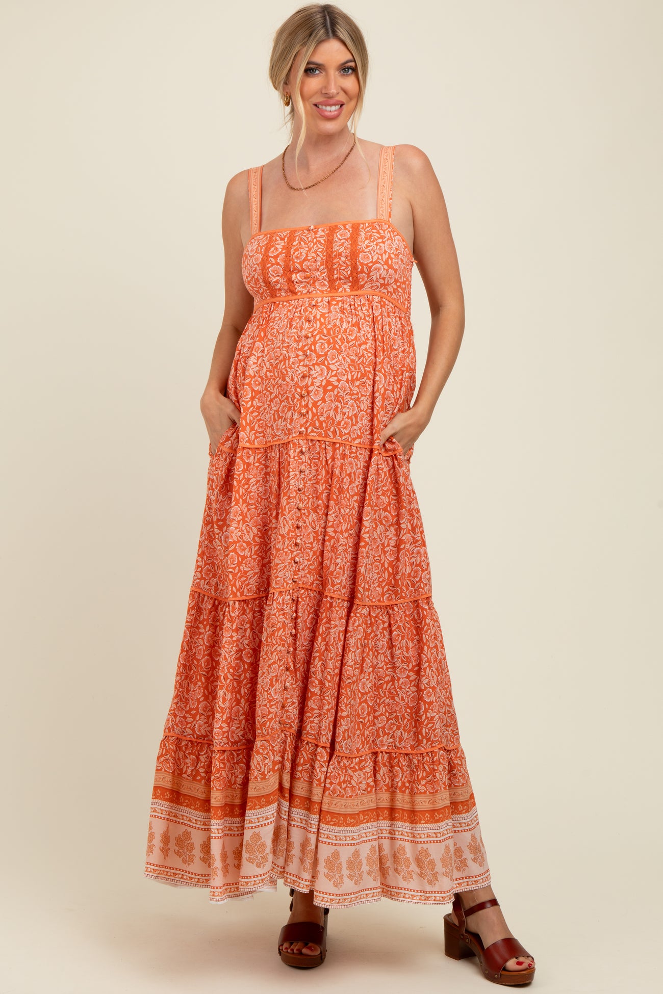 woman wearing an orange patterned maternity maxi dress with tiered details and pockets, smiling while showcasing her baby bump