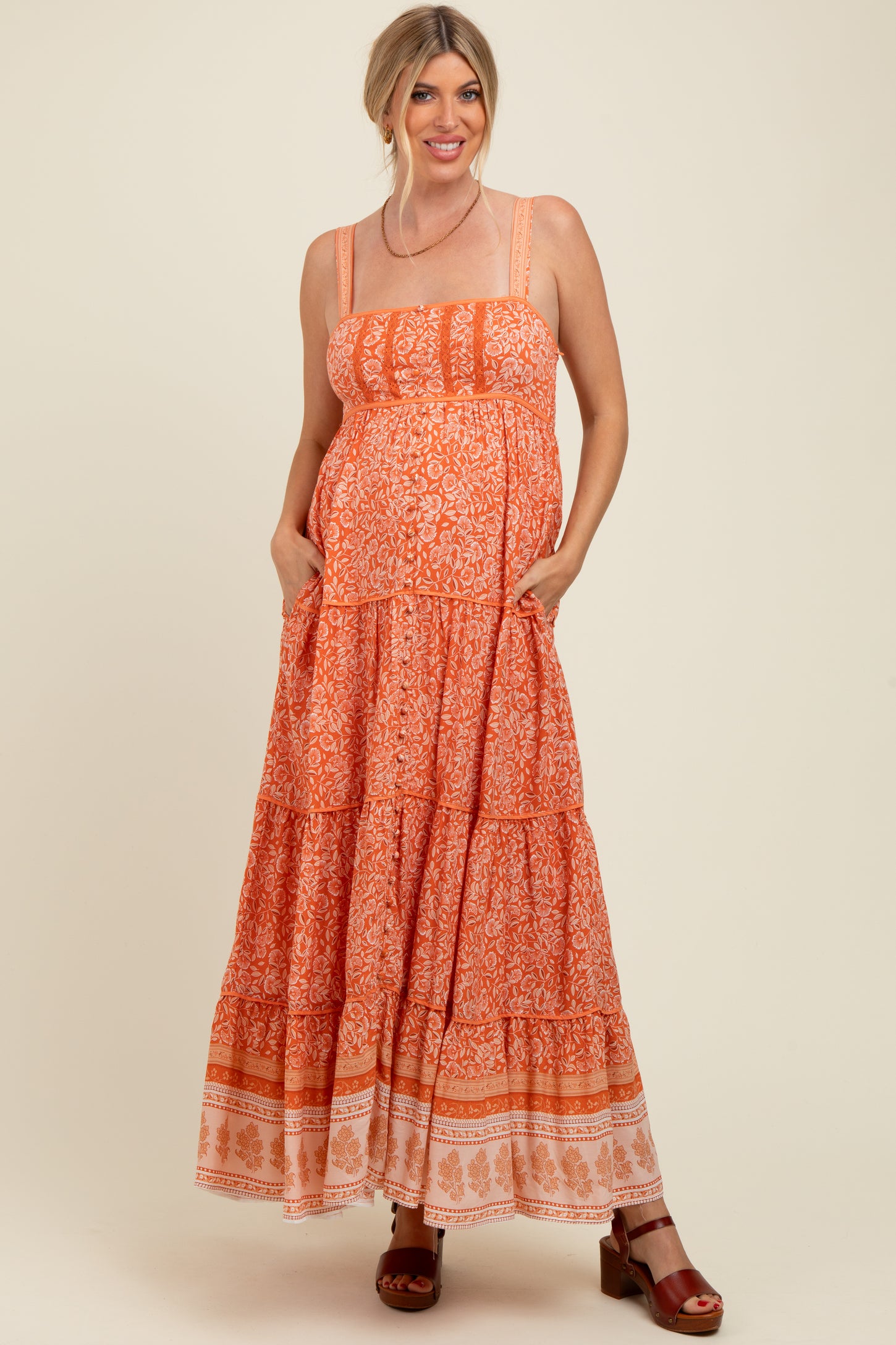 woman wearing an orange patterned maternity maxi dress with tiered details and pockets, smiling while showcasing her baby bump