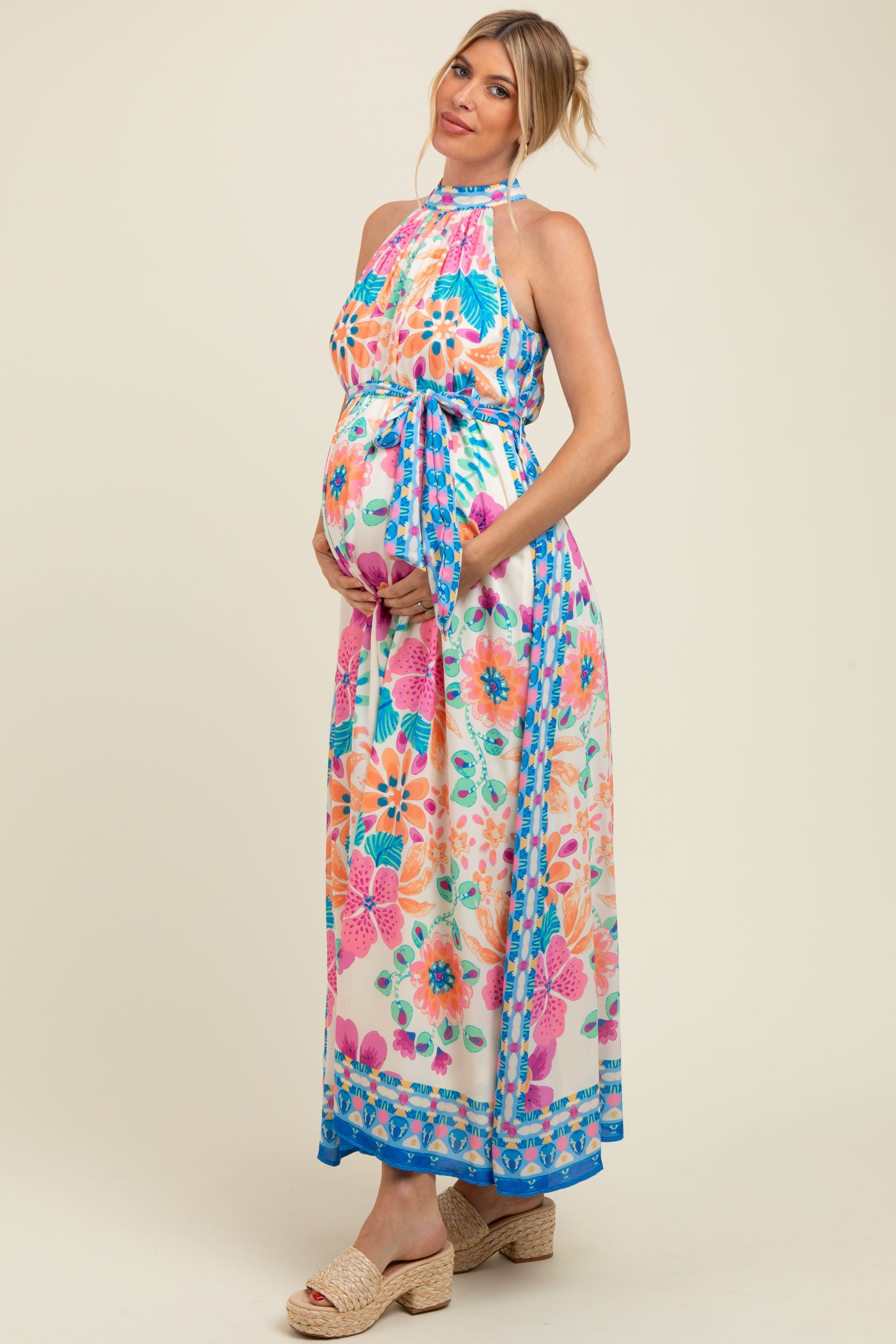 woman wearing a vibrant floral printed maternity maxi dress with halter neck and tie waist, showing her baby bump