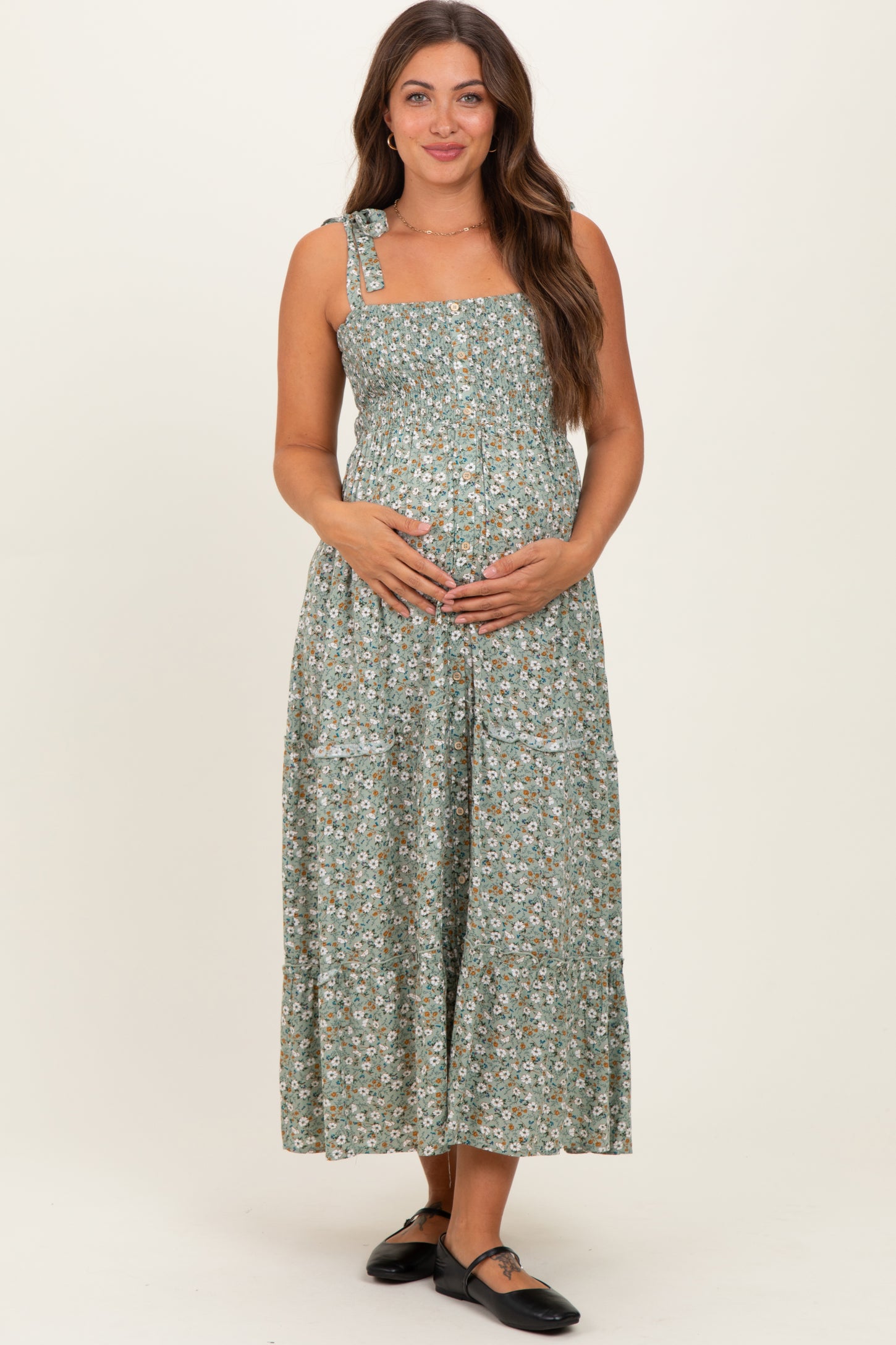 woman wearing a green floral maternity dress with adjustable straps and holding her baby bump