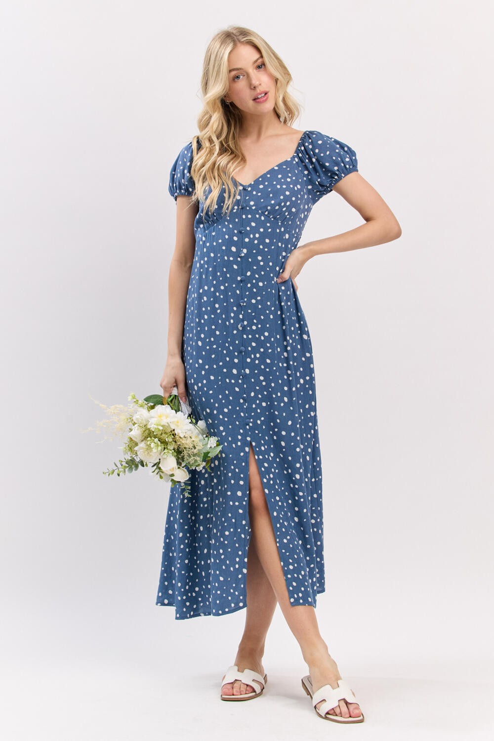 woman wearing a blue polka dot maxi dress with puff sleeves and a front slit, holding a bouquet of flowers