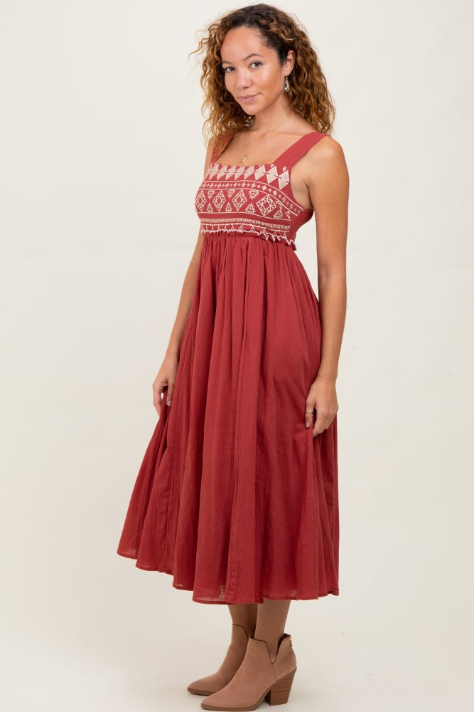 woman wearing a rust red midi dress with embroidered bodice and wide straps, paired with tan ankle boots