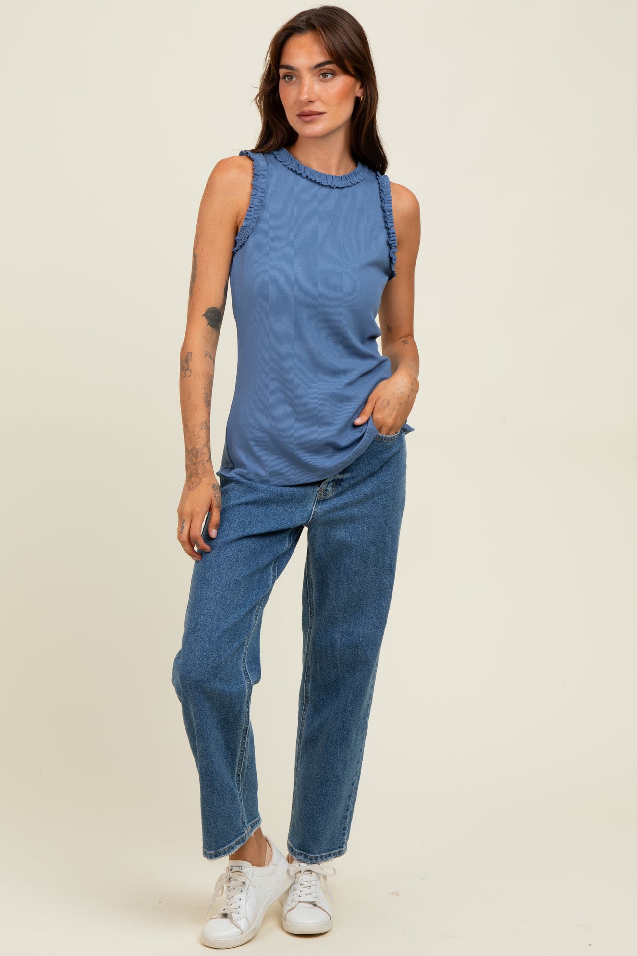 woman wearing a blue sleeveless top with ruffle details and high neckline, paired with high-waisted blue jeans and white sneakers