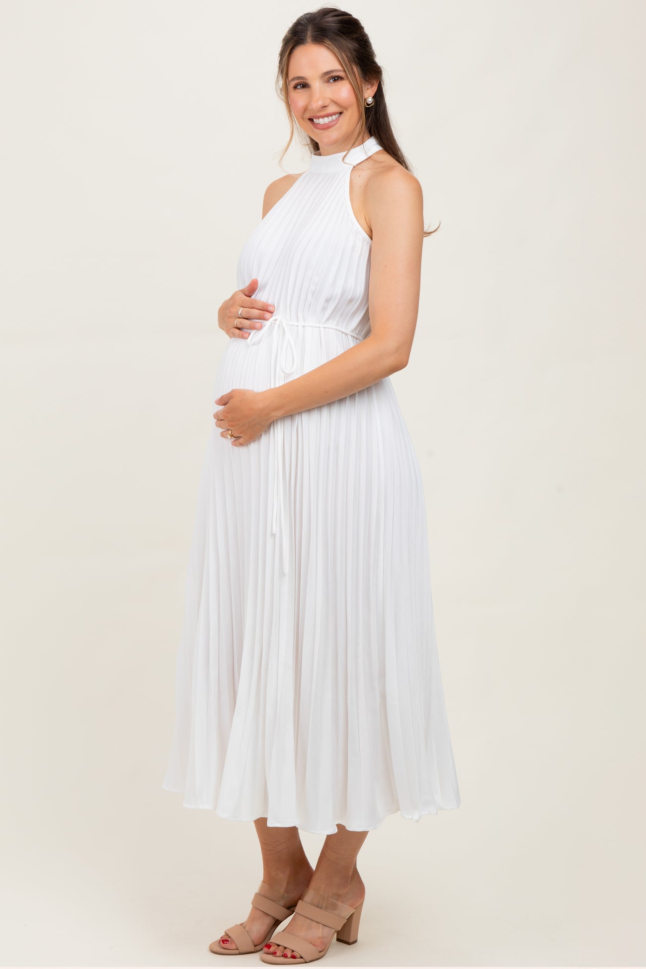 woman wearing a white pleated maternity dress with a high neckline and holding her baby bump