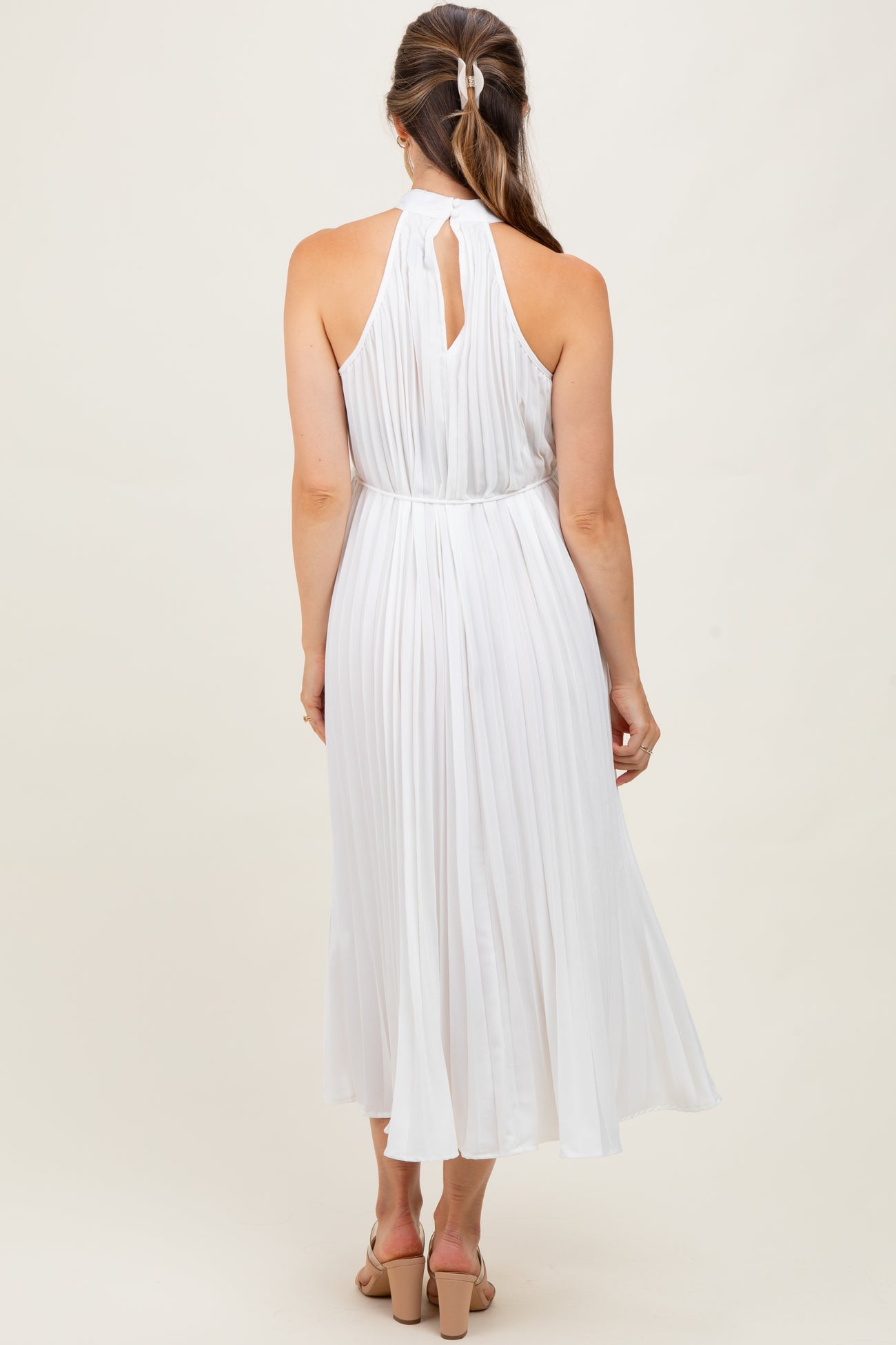 woman wearing a white pleated maternity dress with a halter neck and open back, showing a flowing skirt