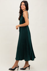 woman wearing a dark green pleated maxi dress with straps and high heels, standing sideways against a neutral background