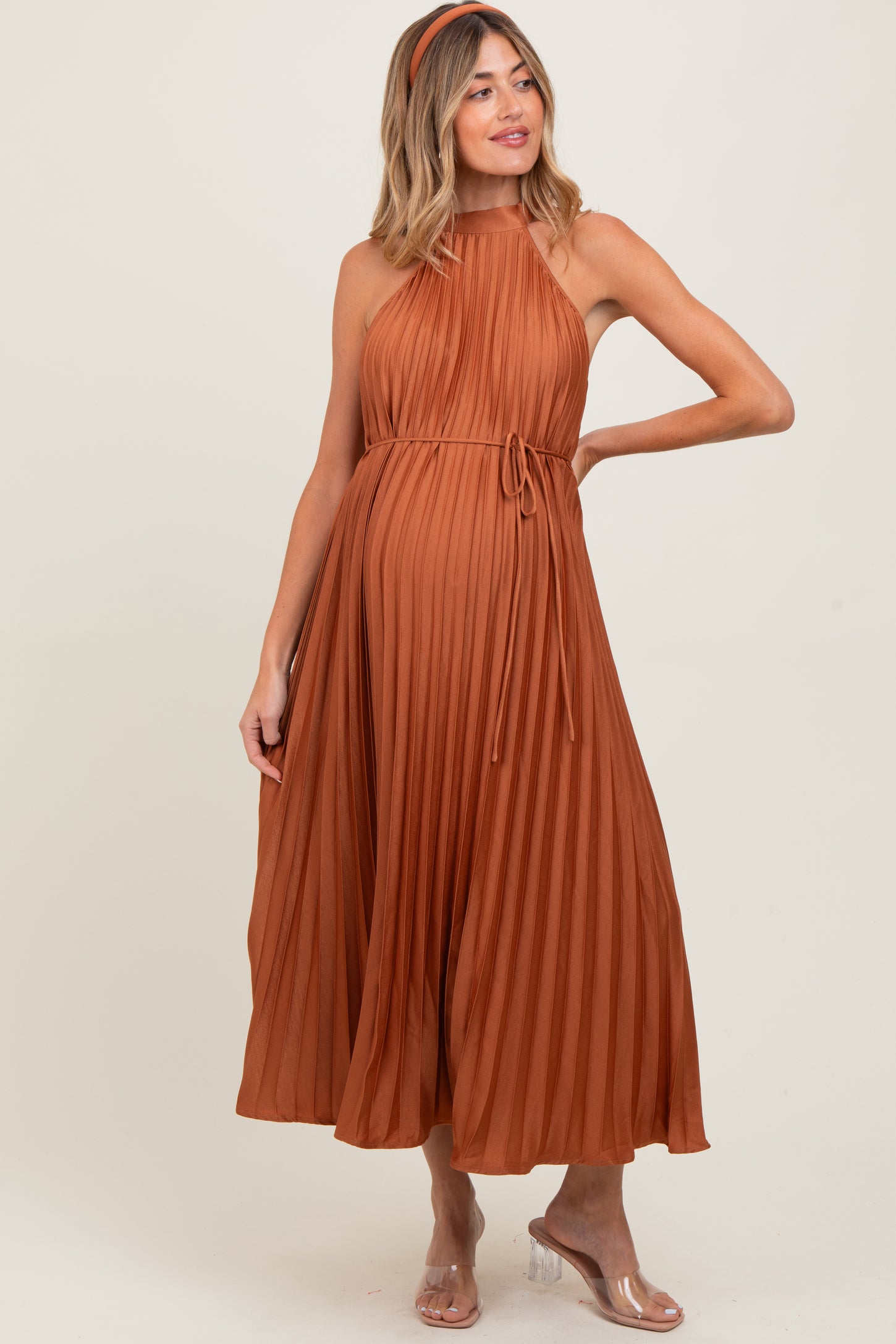 woman wearing a rust pleated maternity dress with a tie waist and halter neckline, showing her baby bump