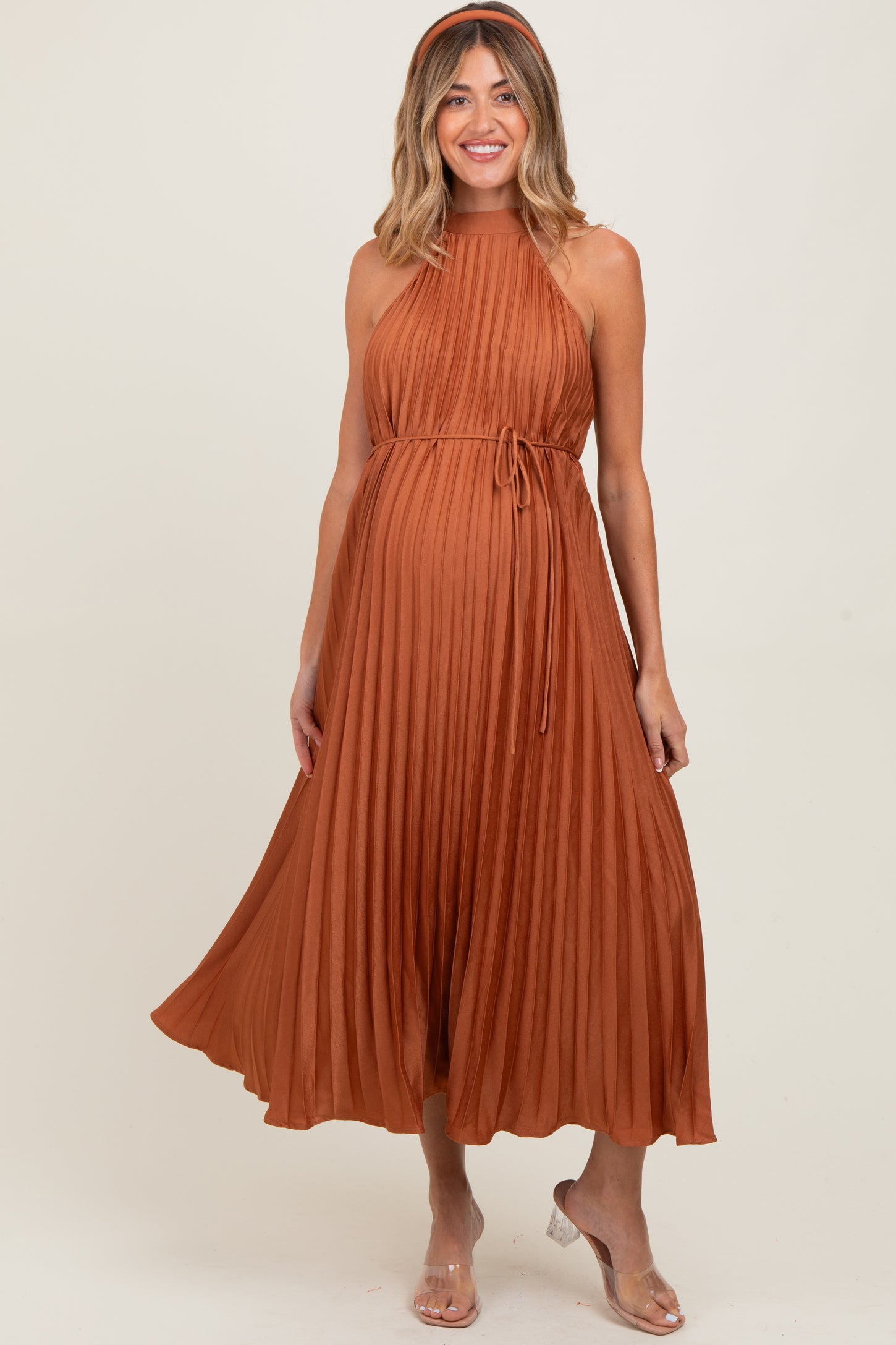 woman wearing a rust pleated maternity dress with a halter neck and a tied waist, showcasing her baby bump