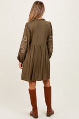 woman wearing a dark olive green maternity dress with embroidered long sleeves and a flowy silhouette, standing with her back to the camera