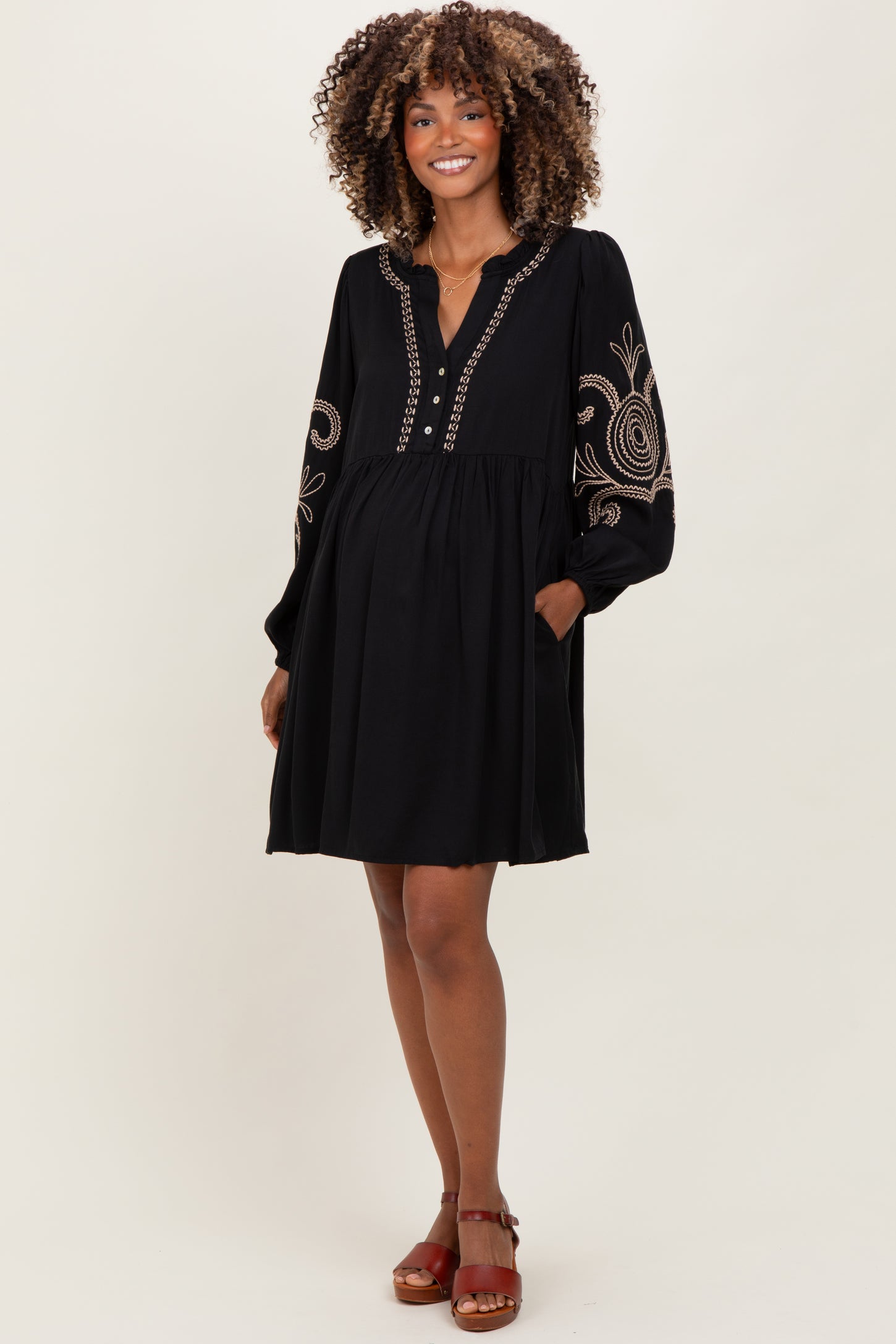woman wearing a black maternity dress with embroidered details and long sleeves, standing with hands in pockets and showing baby bump